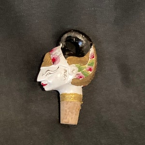 May include: A white ceramic bottle stopper featuring a painted design of a woman's face with floral accents. The stopper is topped with a cork.