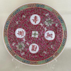 May include: A round decorative plate with a deep red background and a patterned border. The plate features four white circles with Chinese characters and floral accents. The border is a mix of blue, yellow, and green geometric designs.