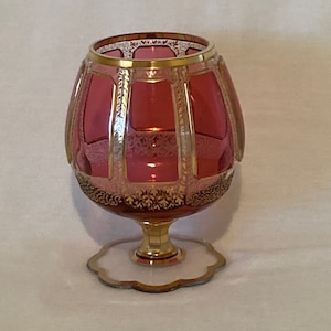 Puede incluir: Una decantador de vidrio vintage con cuerpo rosa y ribetes dorados. La decantador tiene una base ancha y un cuerpo redondo con un patrón decorativo. La decantador está sobre una base blanca con ribetes dorados.