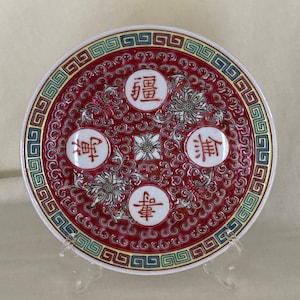 May include: A red and white ceramic plate with a decorative border and Chinese characters. The characters are "福" (fú), "祿" (lù), "寿" (shòu), and "禧" (xǐ).