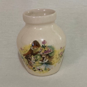 Może przedstawiać: Mały, biały ceramiczny wazon z kwiatowym wzorem przedstawiającym dwoje dzieci w ogrodzie. Wazon ma zaokrąglone ciało i lekko rozszerzoną krawędź.