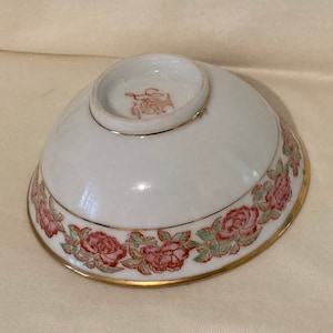 Könnte beinhalten: Weiße Porzellanschale mit goldfarbenem Rand und einem Blumendesign mit rosa und grünen Blumen. Der Boden der Schale trägt den Text "Saint" in Rot.