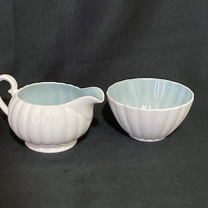 May include: A white creamer and sugar bowl set with a light blue interior. The creamer has a handle and a rounded shape. The sugar bowl is round and has a fluted edge.