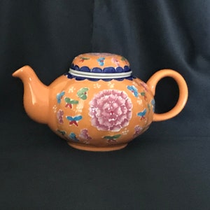 Könnte beinhalten: Orangefarbene Keramik-Teekanne mit Deckel, verziert mit einer großen rosa Pfingstrose und kleineren Blumen- und Schmetterlingsmotiven. Der Deckel hat einen blau-weißen Rand. Die Teekanne hat einen gebogenen Ausguss und einen runden Griff.