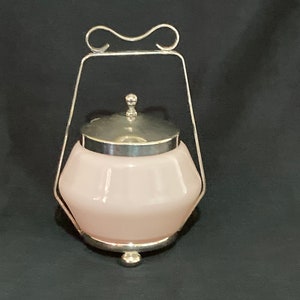 Könnte beinhalten: Eine rosa Glasdose mit silbernem Deckel und einem silbernen Drahtgriff. Die Dose hat ein dekoratives, facettiertes Design und steht auf einem silbernen Sockel.