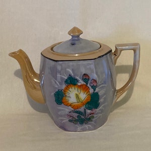 Puede incluir: Una tetera morada con ribete dorado y un diseño floral. La tetera tiene una tapa con la parte superior puntiaguda y un asa en el lateral.