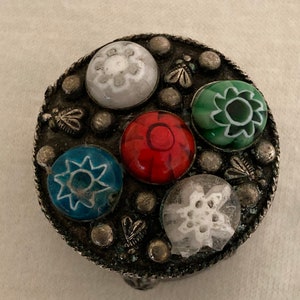 Puede incluir: Una pequeña caja redonda de plata con seis piedras de vidrio de colores engastadas en la parte superior. Las piedras son rojas, verdes, azules y blancas con diseños florales.