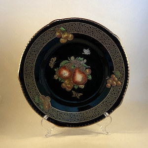 May include: A black decorative plate with a gold rim and a detailed fruit and butterfly design. The plate features a central arrangement of red and yellow fruits, surrounded by grapes and butterflies. The rim has a gold dotted pattern.