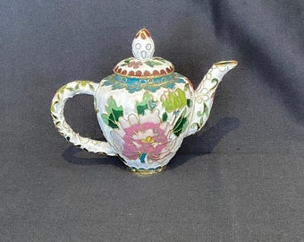 May include: A small white teapot with a floral design. The teapot has a lid with a gold knob and is decorated with pink, green, and blue enamel.