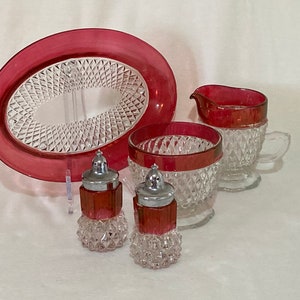 May include: A vintage cranberry glass serving set with diamond-cut detailing. The set includes an oval platter, a creamer, a sugar bowl, and two salt and pepper shakers. The rims of the pieces are trimmed in cranberry.