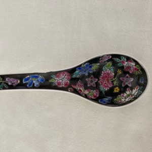 May include: A black ceramic soup spoon with a floral design in shades of pink, blue, green, and yellow. The spoon is decorated with a variety of flowers and leaves.
