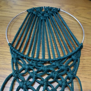May include: A dark green macrame wall hanging with a circular metal hoop and long fringe. The macrame is knotted in a geometric pattern.