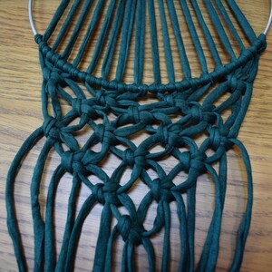 K&ouml;nnte beinhalten: Ein gr&uuml;ner Makramee-Wandbehang mit einem runden Metallring oben. Der Makramee ist aus dicken, gekn&uuml;pften Schn&uuml;ren gefertigt.