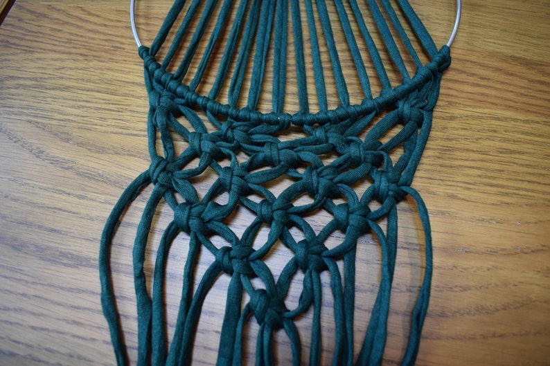 K&ouml;nnte beinhalten: Ein gr&uuml;ner Makramee-Wandbehang mit einem runden Metallring oben. Der Makramee ist aus dicken, gekn&uuml;pften Schn&uuml;ren gefertigt.