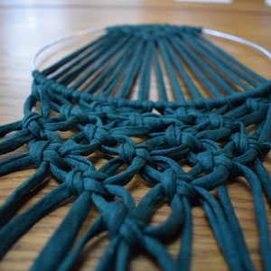 K&ouml;nnte beinhalten: Ein tealblauer Makramee-Wandbehang mit einem runden Metallring und langen Fransen.