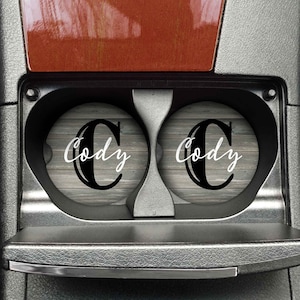 May include: Two gray and white wood-grain car coasters with the name "Cody" in a black and white font.