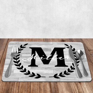 May include: A black and white personalized placemat with a woodgrain pattern. The placemat features a large black letter "M" with a black wreath and the name "Miller" written in white cursive script.