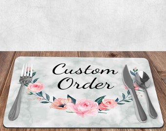 May include: A rectangular placemat with a marble-like background and the words "Custom Order" in elegant script. The placemat features a floral design with pink roses and green leaves. A fork, knife, and spoon are placed on the mat.