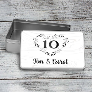 May include: A white metal tin with a black and white design. The design features a heart-shaped wreath with the number 10 inside and the text "Tim & Carol".