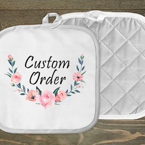May include: Two white pot holders. One features the text "Custom Order" surrounded by a floral wreath with pink flowers and green leaves. The other has a quilted pattern. Both have a white trim and a loop for hanging.