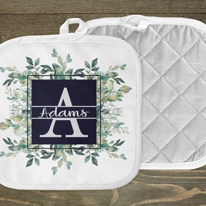 May include: White pot holder with a green leafy design and a navy blue square with the letter A and the name Adams in white text.