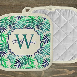 May include: A pair of white quilted pot holders with a green and blue leafy pattern. One pot holder has a monogrammed initial "W" with the name "Williams" below it.