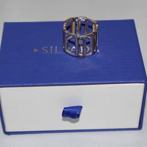 May include: A silver-colored ring with a cage-like design sits atop a blue and white jewelry box. The ring has a geometric, open structure. The box has a pull tab made of blue ribbon and the word "SIL" is visible on the box.