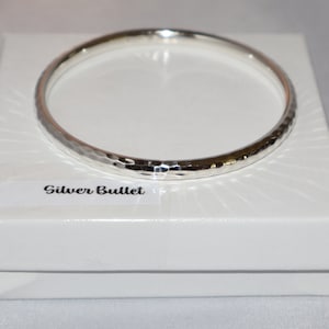 May include: A silver bangle bracelet with a hammered finish. The bracelet is displayed in a white box with the words "Silver Bullet" printed on a label.
