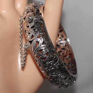 May include: A silver-toned bangle bracelet with intricate floral and scrollwork designs. The hinged bracelet features a safety chain and clasp. The openwork design allows light to pass through, creating a delicate and ornate look.