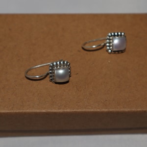 May include: A pair of silver earrings with a white pearl in a square setting. The earrings are on a brown background.