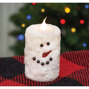 May include: A white snowman candle with a red nose and black eyes. The candle is lit and has a warm glow. The candle is sitting on a red and black plaid surface.