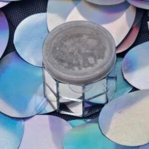 Puede incluir: Un pequeño recipiente redondo de plata con una tapa de espejo. El recipiente está sobre un fondo azul y plateado.