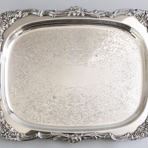 May include: A rectangular silver serving tray with ornate scrollwork and handles. The tray has a raised edge and a decorative border. The surface is engraved with a floral pattern. The tray measures approximately 20 inches long and 14 inches wide.
