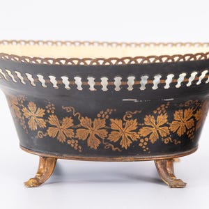 May include: An oval metal bowl with a black exterior featuring gold-colored grape leaf and vine patterns. The bowl has a scalloped rim, a perforated upper border, two small side ring handles, and stands on three curved, gold-colored metal feet.