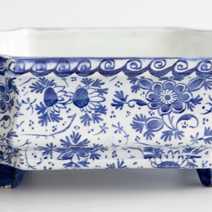 May include: An octagonal ceramic bowl featuring a white base with intricate blue floral patterns. The bowl has a scalloped rim with a wave-like border, two sculpted lion head handles on opposite sides, and stands on four small, sturdy blue feet.