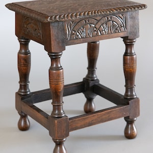 May include: A small, antique wooden stool with a square top and carved floral details. The dark brown stool has turned legs and a lower frame for stability. The top is approximately 12 inches square.
