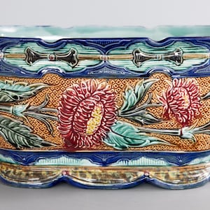 May include: Rectangular ceramic planter with a detailed floral design. The planter features a light blue interior, a dark blue border, and a textured orange body with red and yellow flowers. Gold lion head handles are on each side.