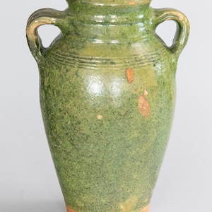 May include: A green ceramic vase with two handles. The vase has a speckled texture and a light brown base. The top rim and handles are a darker shade of green. The vase is a decorative item.