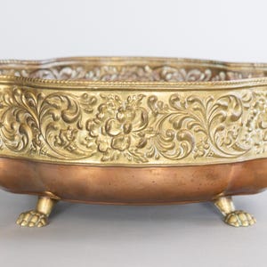 May include: An antique brass and copper decorative bowl. The bowl has a detailed floral pattern around the rim and is supported by three claw feet. The handles are ornate and add to the vintage aesthetic.
