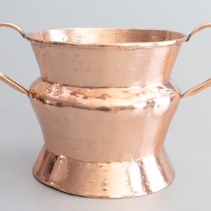May include: A copper-colored vessel with two curved handles. The hammered metal has a warm, reflective sheen. The vessel has a wide, open top and a flared base. The overall shape is reminiscent of a chalice or decorative bowl.
