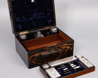May include: An antique wooden box with a black and brown woodgrain finish. The box is open and contains a variety of silver-plated grooming tools, including a comb, tweezers, and a nail file. The box is lined with blue velvet.