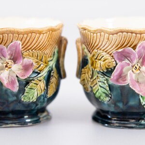 May include: A pair of ceramic planters featuring a deep teal glazed body with a scalloped tan rim. Each planter is decorated with three-dimensional pink flowers and green leaves applied to the exterior, along with small side handles.