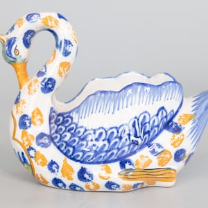 May include: A white ceramic swan planter with blue and yellow painted details. The swan has a wide, open beak and a raised tail. The planter is decorated with a blue and white floral pattern and yellow dots.