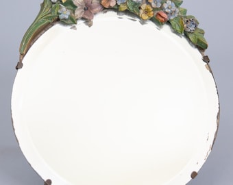 Op de afbeelding: Een ronde spiegel met een witte lijst en een bloemmotief rond de rand. De bloemen zijn geschilderd in tinten roze, geel, blauw en groen.