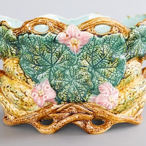 May include: A ceramic planter with a botanical design. The planter features green leaves, pink flowers, and brown branches. It has a scalloped rim and two handles. The planter is approximately 12 inches wide and 6 inches tall.