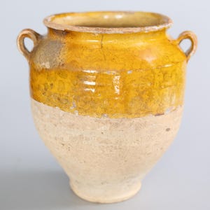 May include: A two-toned ceramic pot with a yellow upper half and a cream-colored lower half. The pot has two small loop handles and a slightly flared rim. The yellow section has a glossy finish, while the cream section has a matte finish.