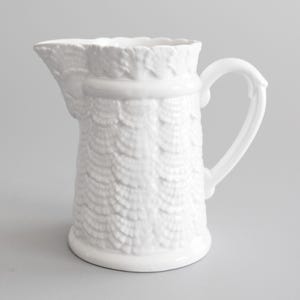 May include: A white ceramic pitcher with a textured, shell-like pattern. The pitcher has a curved handle and a spout for pouring. The rim of the pitcher has a scalloped design. This decorative pitcher is suitable for serving beverages.