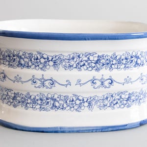 May include: White ceramic planter with blue floral and scrollwork designs. The planter has a blue rim and two handles. The floral pattern is repeated in two bands around the planter's body. A decorative piece for home decor.