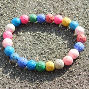May include: A colorful bracelet composed of round, porous beads in various colors, including pink, blue, green, and yellow. The beads are arranged in a circular pattern, resting on a textured surface.