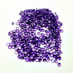 May include: A scattering of small, round, faceted amethyst gemstones. The gems are a deep purple hue and are spread across a white surface. The gemstones vary in size and shape, reflecting light.